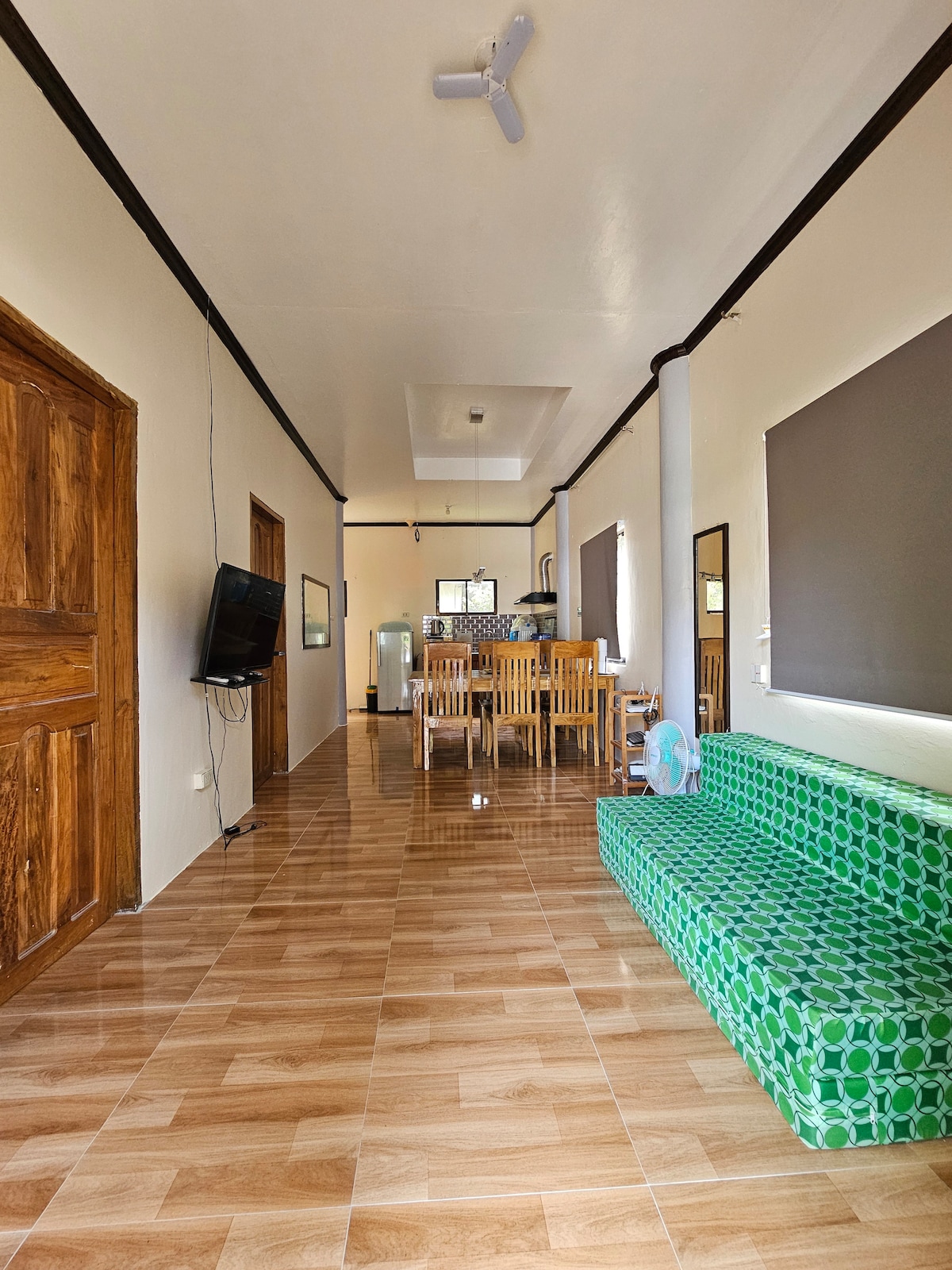 A spacious hallway leads towards a well-lit kitchen and dining area. A green patterned couch is positioned along one wall, while a wall-mounted television is visible opposite. Natural light floods in through large windows, complementing the light-colored walls and tile flooring.