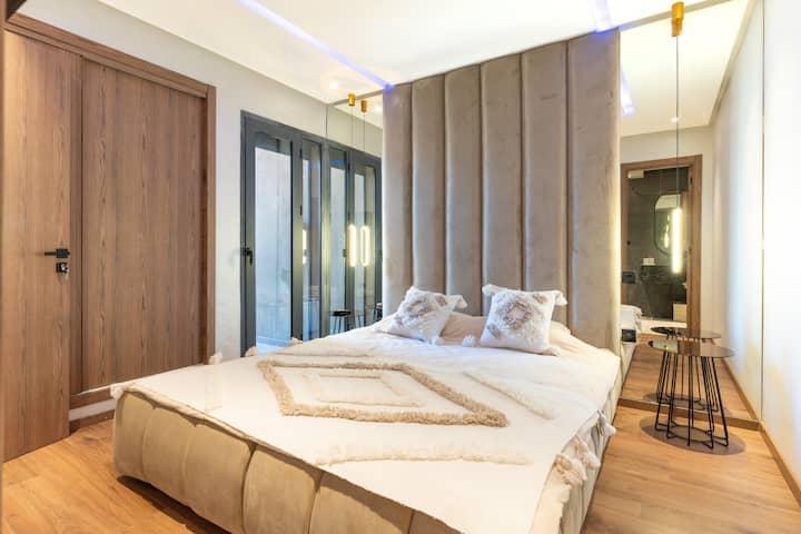 优雅的卧室，带软垫床头板、吊灯和温馨的氛围。 舒适的床、壁挂式电视和独立套内卫生间，绝对舒适。