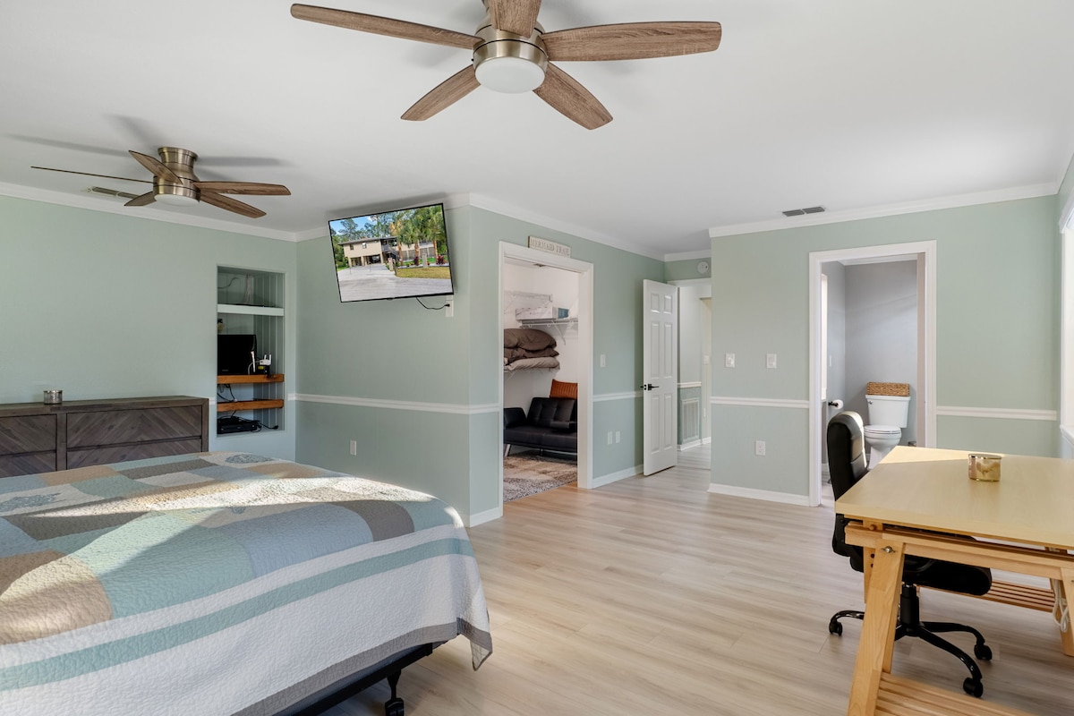 غرفة نوم رئيسية واسعة مع جدران خشبية ساحلية ناعمة، وأرضيات فاخرة من ألواح الفينيل، وسرير كينج، ومراوح سقف مزدوجة، وتلفزيون مثبت على الحائط، وركن مكتب منزلي، ووصول مباشر إلى خزانة ملابس وحمام داخلي.