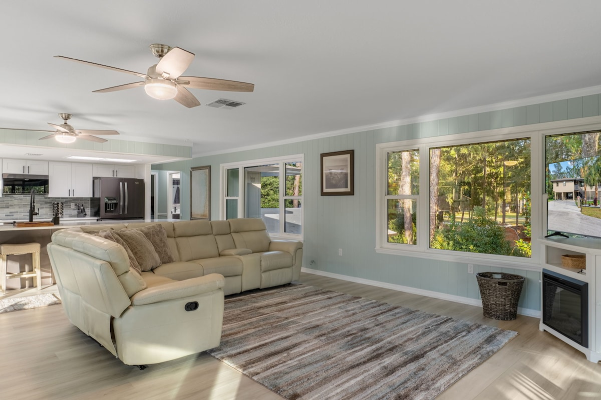غرفة معيشة مشرقة ذات مفهوم مفتوح مع جدران شيبلاب ساحلية وأرضيات فينيل فاخرة وأريكة بيج قطاعية مريحة وإضاءة طبيعية وفيرة وتدفق سلس إلى المطبخ وغرفة التشمس. ملاذ جيد التهوية ومريح.