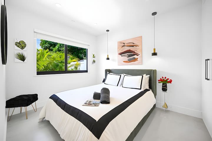 Dormitorio minimalista con ropa de cama acogedora, iluminación moderna, arte mural y ventana con vistas al jardín.