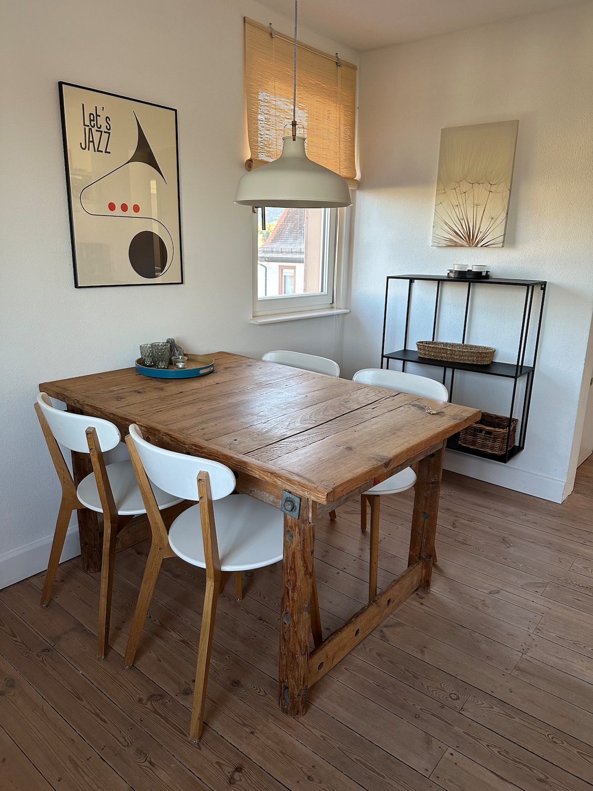 A simple dining area is featured with a wooden table surrounded by four white chairs. A pendant light hangs above the table, illuminating the space. A small shelving unit in the corner holds decorative items and a basket. Natural light comes through the window.