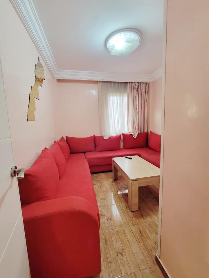 تم تحويل الغرفة الثانية إلى غرفة معيشة بها ثلاث أرائك حمراء وتلفزيون مثبت على الحائط. مساحة مشرقة وعملية في شقة للإيجار في دار بوعزة، وهي مثالية للاسترخاء أو العمل.