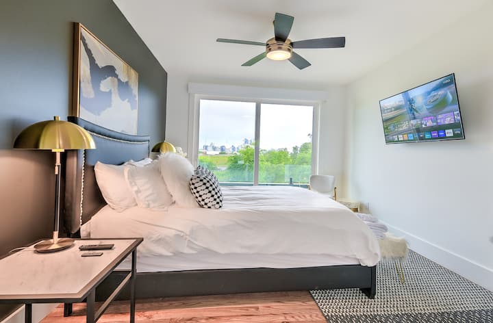 Bright, stylish bedroom with a plush king bed, modern decor, and large windows offering city views. Features crisp white linens, ceiling fan, smart TV, cozy seating, and warm wood floors—perfect for relaxing after a day exploring the city.