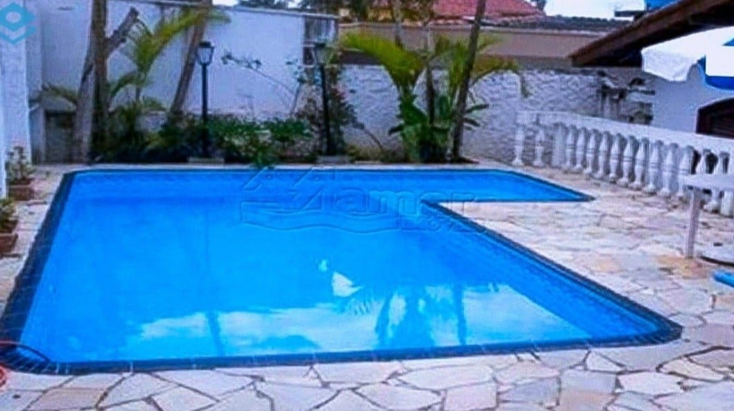 A rectangular pool is surrounded by a paved area, featuring a light-colored stone finish. Lush green plants are positioned near the pool, providing a natural touch. The area appears well-maintained, inviting relaxation and leisure.