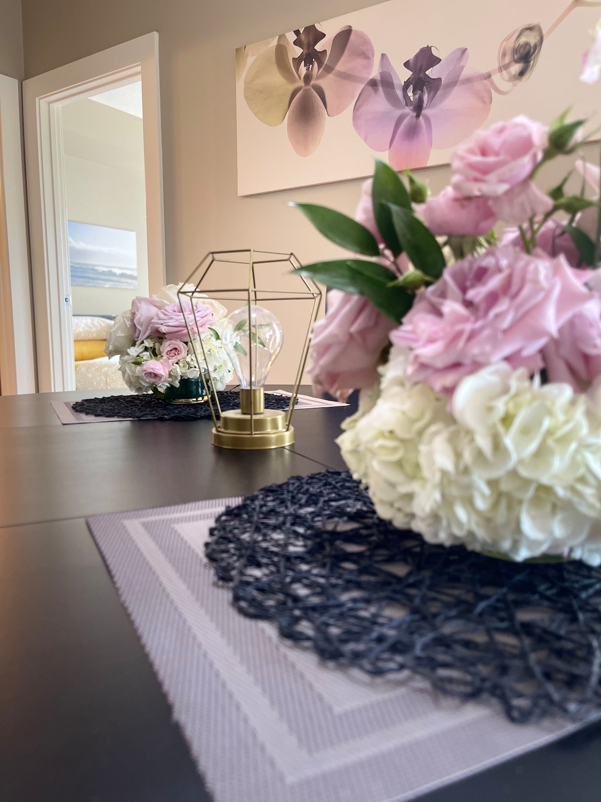A dining table is adorned with a floral arrangement featuring pink roses and white hydrangeas. A golden geometric vase adds a decorative touch. The table setting includes dark woven placemats, while soft artwork depicting orchids hangs on the wall in the background.