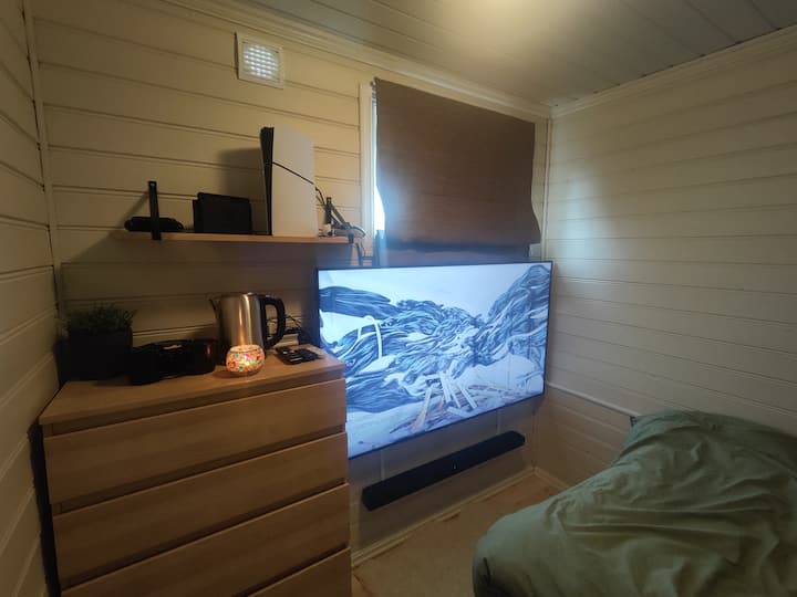 Aquí puedes jugar un poco a la Nintendo Switch o a la PlayStation 5 (solo hay un control y un cable HDMI para cada una de las consolas de videojuegos). Televisor de 75”. Hay una barra de sonido conectada con un subwoofer debajo de la cama y Google Chromecast. Los tres cajones superiores están vacíos.