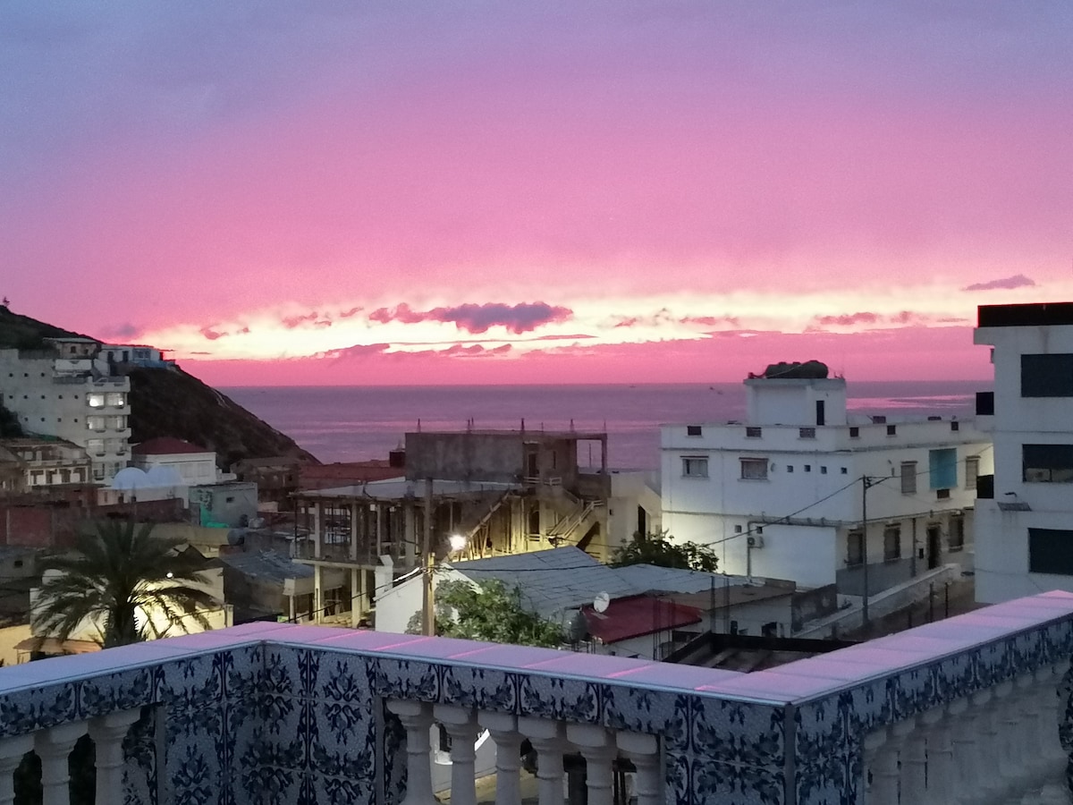 A vibrant sunset fills the sky with hues of pink and purple, creating a striking backdrop over the coastline. The view extends to buildings and rooftops below, with a palm tree partially visible in the foreground, hinting at the lively atmosphere of the area.