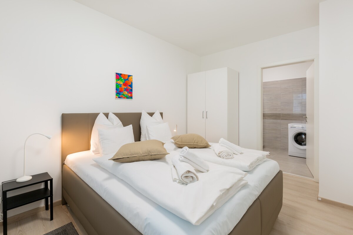 Spálňa má pohodlnú manželskú posteľ, mäkkú posteľnú bielizeň a upokojujúci dekor, čo vytvára ideálne prostredie pre pokojný spánok. Veľké okná poskytujú prirodzené svetlo, zatiaľ čo zatemňovacie závesy zaisťujú súkromie a pohodlie.