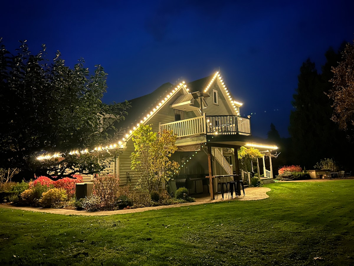 The exterior of the home is illuminated by warm string lights against a darkening sky. A covered balcony extends from the second floor, while the surrounding yard features well-maintained greenery and colorful plants, creating a tranquil ambiance.