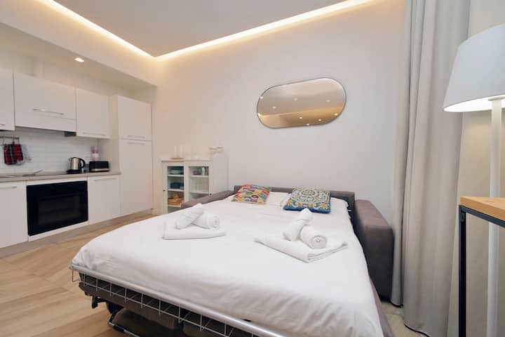 Moderan studio apartman s elegantnom LED stropnom rasvjetom, udobnim bračnim krevetom s čistom posteljinom, kompaktnom čajnom kuhinjom s bijelim ormarićima, modernim ovalnim ogledalom, podovima od tvrdog drva s uzorkom riblje kosti i učinkovitim dizajnom otvorenog plana savršenim za boravak u Rimu