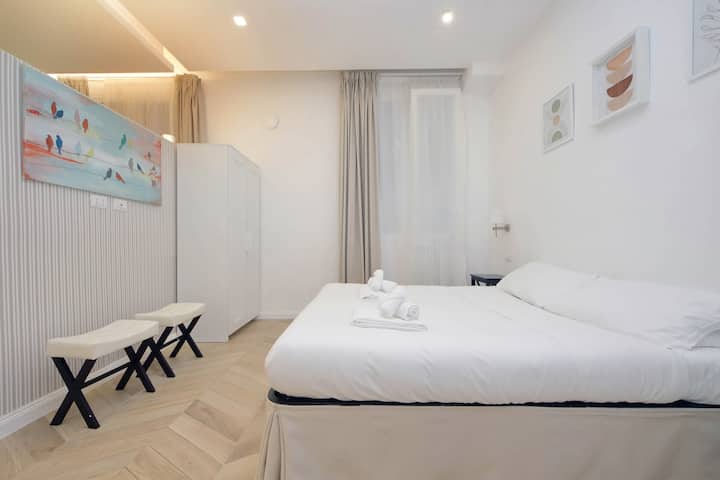 Svijetla spavaća soba s bračnim krevetom, bijelom posteljinom, umjetničkim radovima i podom s uzorkom riblje kosti. Minimalistički uređenje i prirodno svjetlo stvaraju ugodan i elegantan prostor, savršen za opuštajući i elegantan boravak.