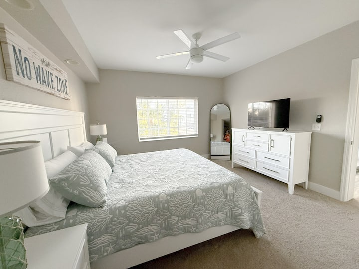 Dormitorio principal tranquilo con ropa de cama con motivos costeros, ventilador de techo y luz natural. Disfrute de una cómoda cama king, un televisor de pantalla plana y una decoración con estilo playero, incluido un letrero de «Zona de no despertar» para crear un ambiente relajado en Fort Myers Beach.