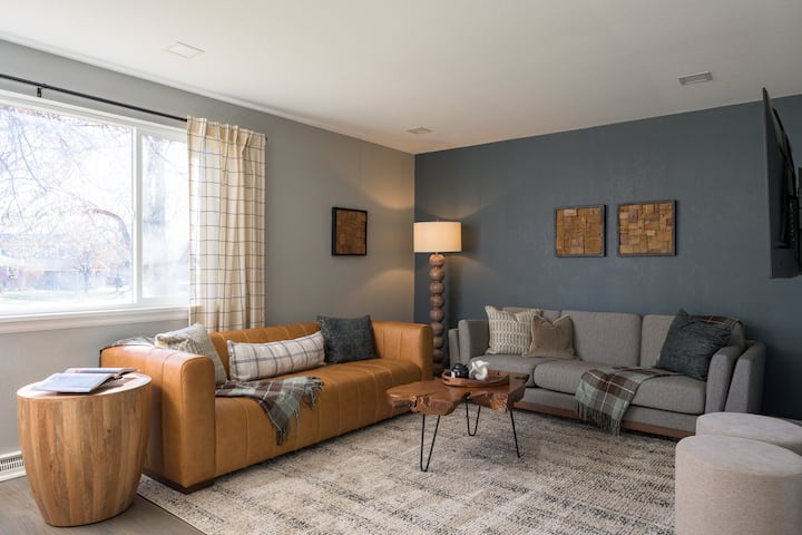 Sala de estar luminosa y elegante con un sofá de cuero, asientos acogedores, iluminación cálida y una mesa de centro única con borde vivo. Los grandes ventanales llenan el espacio de luz natural, perfecto para relajarse después de un día en Denver.