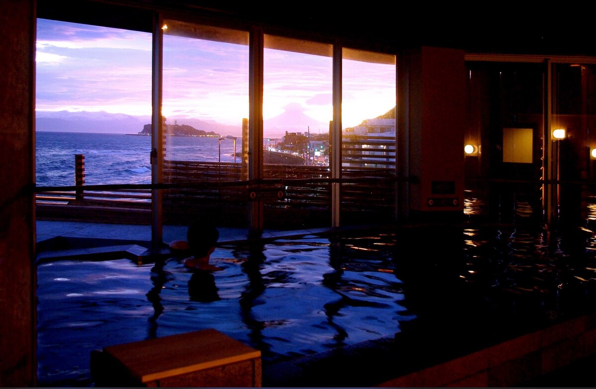 A tranquil bath area is visible, featuring a large indoor pool with calm water reflecting the vibrant colors of the sunset. Floor-to-ceiling windows provide views of the coastline and distant hills, creating a serene backdrop for relaxation.