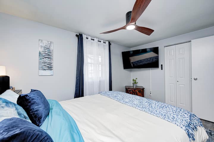 Светла, мирна спална соба со удобен кревет облечен во свежа постелнина, меки сини детали и модерен вентилатор на таванот. Ѕидниот телевизор од 55 инчи овозможува лесно гледање, додека природната светлина, чистите линии и неутралните тонови создаваат релаксирачко прибежиште.