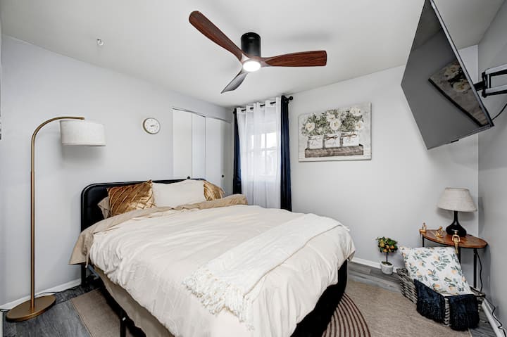 Примамлива спална соба со мек кревет со мека неутрална постелнина и украсни перничиња со текстура. Модерниот тавански вентилатор, телевизорот од 55 инчи монтиран на ѕид и топлото осветлување создаваат опуштена атмосфера, додека чистите линии и едноставниот декор го одржуваат просторот спокоен.