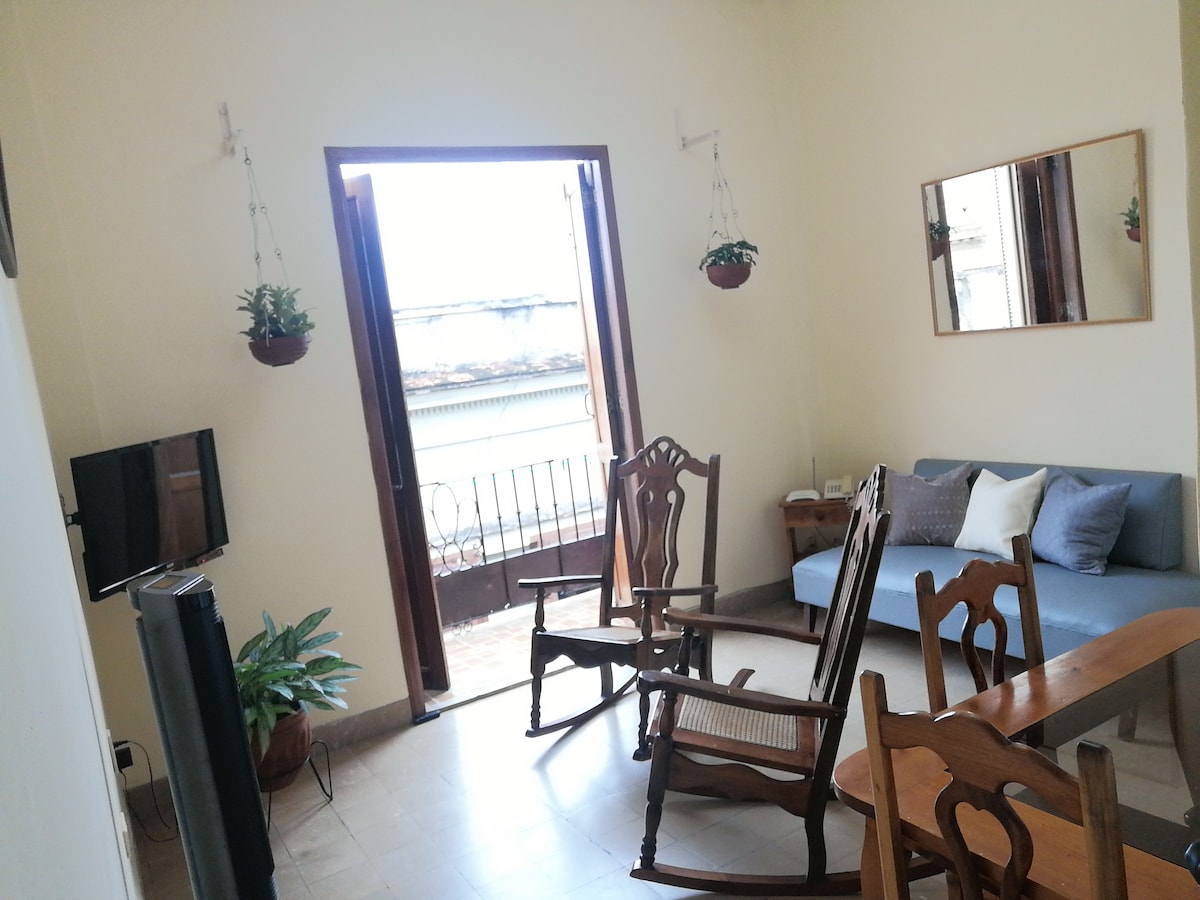 The living area features a combination of seating options including two rocking chairs and a sofa, with natural light flooding in from the open door leading to the balcony. A wooden table and a television are also visible, creating a comfortable space.