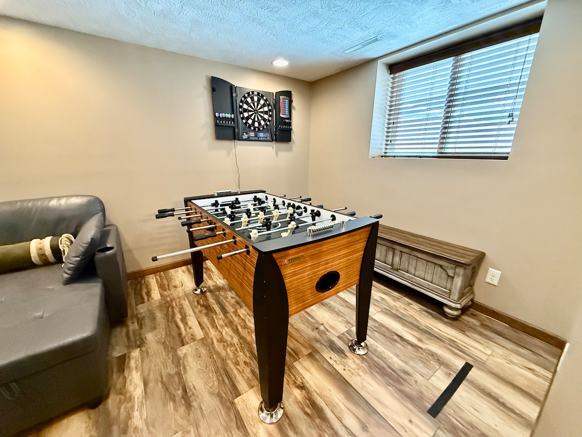 Eneo la chini la kuishi lenye sofa mbili za kulala, meza ya mpira wa meza, ubao wa mchezo wa kurusha vishale, kiti kikubwa cha mfuko wa maharagwe na televisheni janja ya inchi 65. 