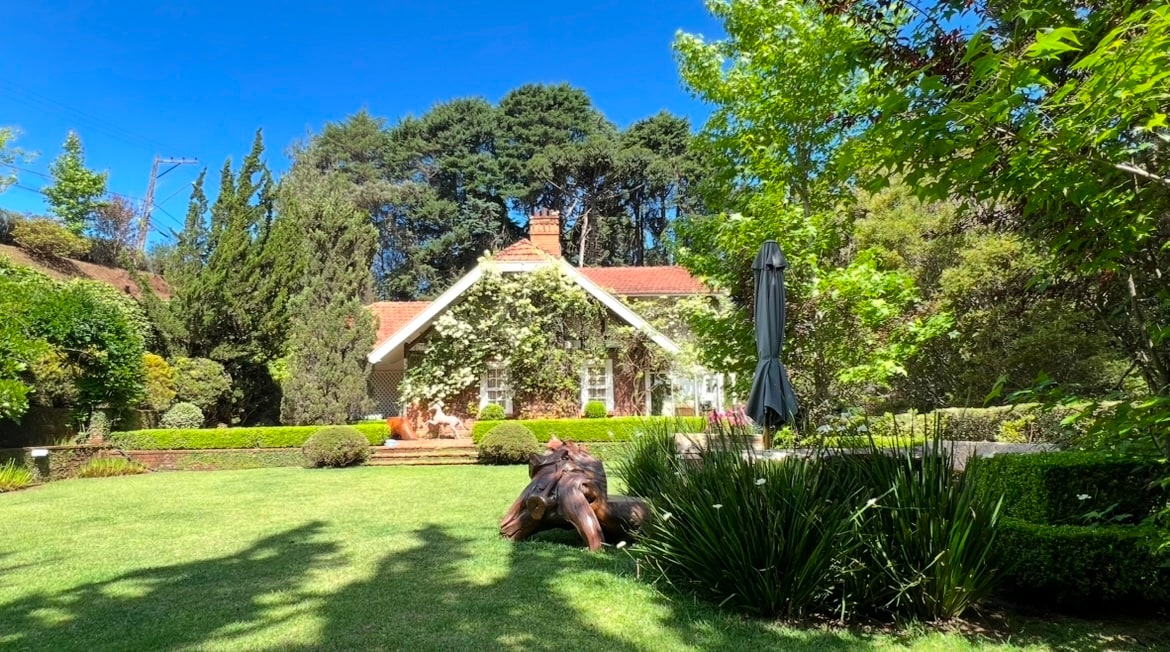A serene garden is showcased, featuring well-manicured lawns and vibrant greenery. The house, framed by trees and shrubs, displays classic architecture. A large sculpture rests on the lawn, adding an artistic touch to the outdoor space.