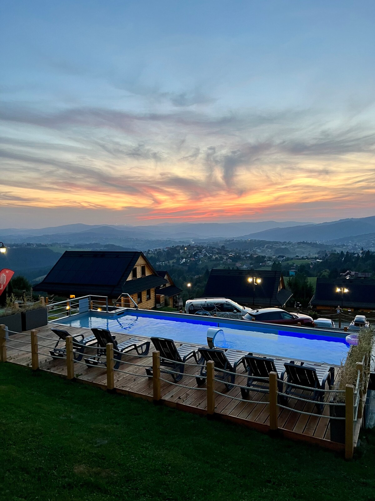 An outdoor area features a swimming pool surrounded by lounge chairs, set against a backdrop of fading sunlight over distant mountains. The wooden deck provides a relaxing space, and atmospheric lights add a gentle glow to the surroundings as day transitions to night.
