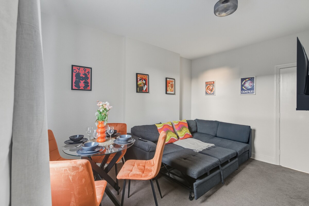 พื้นที่นั่งเล่นทันสมัยสว่างสดใสพร้อมเตียงโซฟาสีเทา เบาะหลากสี โต๊ะรับประทานอาหารทรงกลม เก้าอี้สีส้ม และงานศิลปะติดผนังที่มีสไตล์สร้างความรู้สึกอบอุ่นและร่วมสมัย