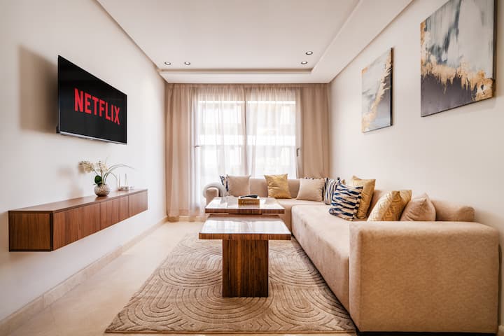 • Svetlá, útulná obývacia izba: veľká béžová pohovka v tvare L, nástenný televízor so službou Netflix (účet pre hostí), drevený konferenčný stolík + hrubý koberec. Veľké okno/dvere s priehľadnými závesmi, čistý elegantný dekor, ideálne na relax alebo prácu.