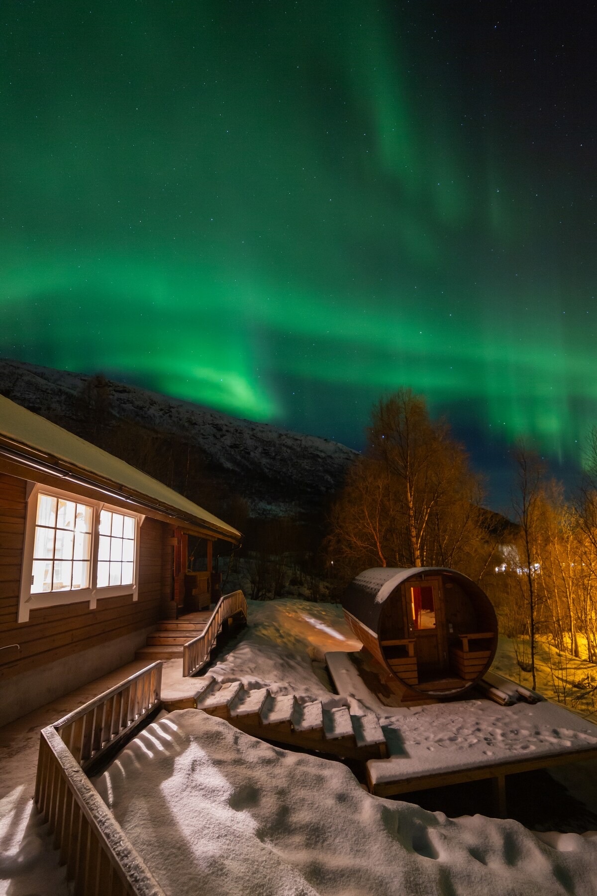 The outdoor area features a cozy barrel sauna alongside a snow-covered pathway leading to a warm cottage. Above, the northern lights create a vibrant green display against a starlit sky, illuminating the surrounding snow and distant mountains.