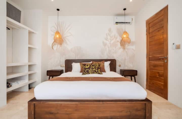Duerme con estilo en este dormitorio sereno con una cama de teca hecha a mano y un mural tropical personalizado. La cálida iluminación de ratán y las sábanas de felpa garantizan una noche de sueño reparador en el paraíso.