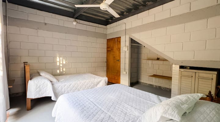 ห้องพักสองเตียงที่เงียบสงบพร้อมเตียงเดี่ยวสองเตียงที่ปูผ้าคลุมสีขาวและพัดลมเพดานทรงพลัง พื้นที่พักผ่อนที่สดชื่นและเรียบง่าย