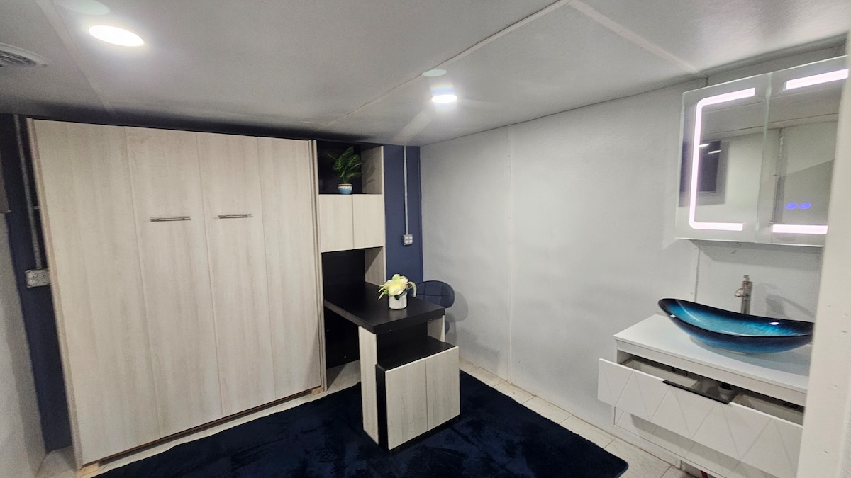 Veľká sklápacia posteľ vo viacúčelovej izbe. Izba má tiež malú kúpeľňu, televízor na stene a stôl so stoličkou.