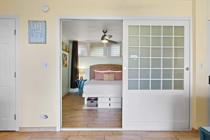 Queen bedroom with wall-mounted TV, luggage rack, and chic barn doors. Dual fans (ceiling & floor oscillating)  keep the space  cool.