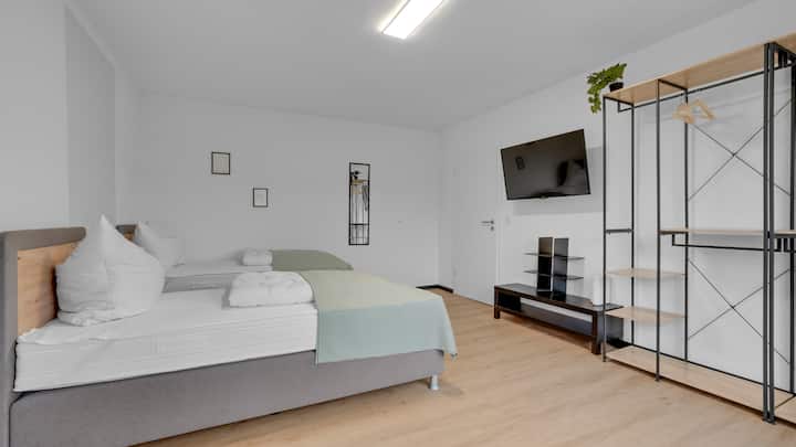 Пространа спална соба со два единечни кревета, светли јоргани и зелен прекривач. Дрвен под, телевизор монтиран на ѕид, отворени метални полици со дрвени подови, суптилна ѕидна декорација и чист, модерен внатрешен дизајн.