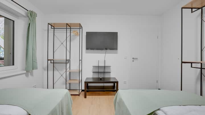 Прекрасна спална соба со два единечни кревета, светлозелени прекривки и дрвен под. Телевизор монтиран на ѕид, отворени метални полици со дрвени подови, прозорци со зелени завеси и чист, функционален мебел.