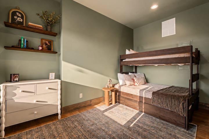 Afternoon light spills across this bunk room's plaid bedding, styled shelves, and fluted dresser topped with a WiFi card — thoughtful, guest-friendly touches throughout.