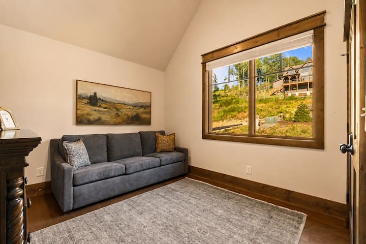 This light-filled bedroom features a sofa bed set beneath a big picture window framing the wooded hillside, with vaulted ceilings letting the sunshine pour in. A bright, comfortable space to rest and take in the mountain scenery.