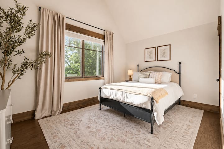 Sunlight pours through rustic wood-trimmed windows into this bright, vaulted bedroom, where soft layered linens on the iron bed frame and a leafy, tree-lined view make for an easy, restful morning.