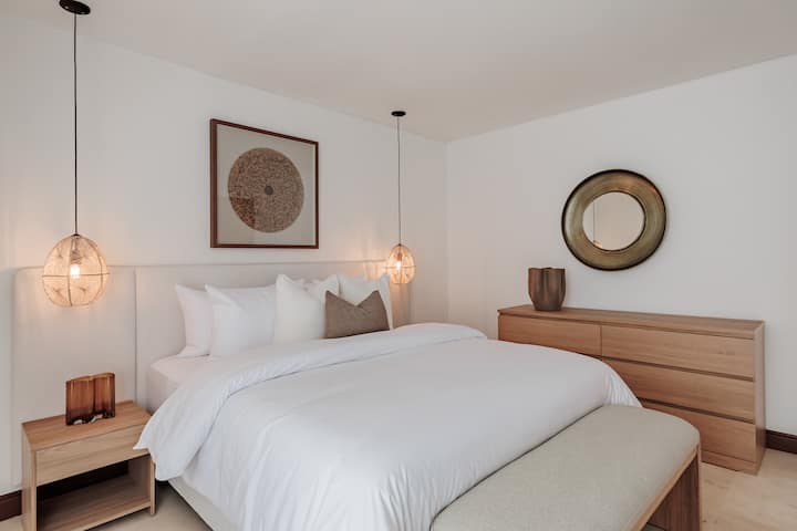 Suite privada #5: Texturas refinadas. Este ángulo resalta la cama tamaño queen y un versátil banco tapizado, perfecto para sentarse o guardar equipaje. Con luces colgantes de paja orgánica, un espacioso vestidor de madera y elegantes suelos de mármol.