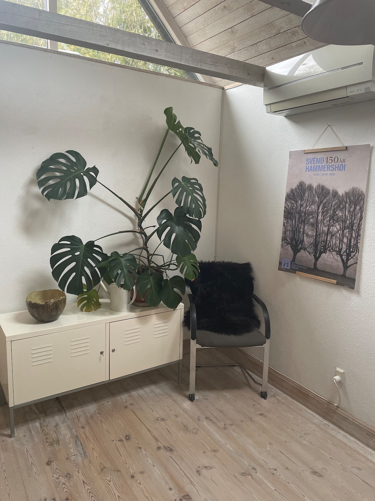 A corner of the room is highlighted by a large green plant, adding a touch of nature. A simple white cabinet sits beneath it, accompanied by a black chair with a plush cushion. A poster featuring trees is displayed on the wall, contributing to the decor.