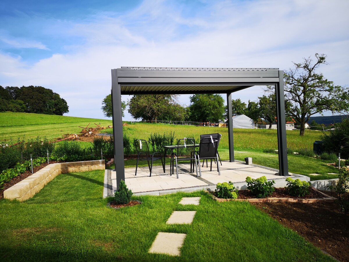 A modern pergola is set on a patio surrounded by well-maintained grass and garden beds. Several chairs are arranged around a table beneath the shelter, with open views of the surrounding green fields and trees in the background. Pathway stones lead towards the pergola.