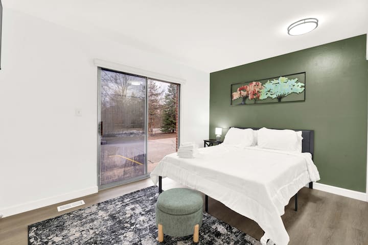 ห้องนอนทันสมัยสว่างสดใสพร้อมเตียงควีนไซส์ ผ้าปูที่นอนสีขาวสดใส และผนังสีเขียวพร้อมงานศิลปะ ประตูกระจกบานเลื่อนนำแสงธรรมชาติเข้ามาและมีทางเข้าสู่ระเบียง สร้างพื้นที่ที่สงบและน่าดึงดูดใจสำหรับการพักผ่อน