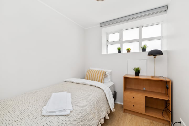 Spálňa s jednolôžkom, čerstvo ustlaná s posteľnou bielizňou, nočnou lampou a malou skrinkou na vaše základné potreby 