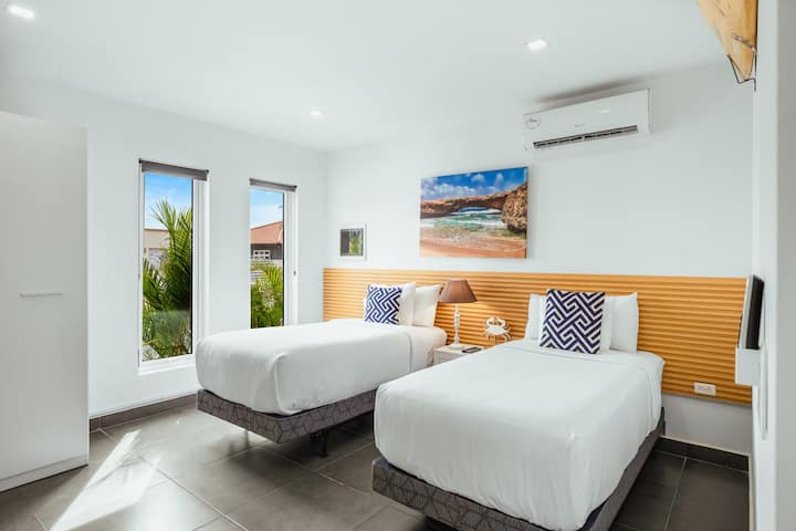 Dormitorio con dos camas individuales bañado por la luz del sol, con sábanas impecables, decoración costera y aire acondicionado refrescante.