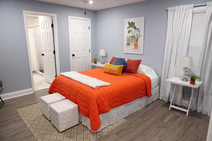 חדר שינה רגוע עם קירות כחולים רכים, שמיכת טלאים כתומה ועיצוב פשוט שמרגיש חם ומאוזן. המיטה מעוצבת לנוחות, עם שידות לילה ותאורה ללילות ובקרים קלים.
