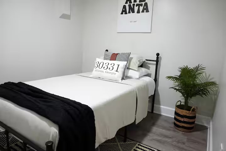 חדר שינה פשוט עם מצעים לבנים נקיים, נגיעות שחורות ועיצוב נקי בכל מקום. המיטה מסודרת בצורה מסודרת למנוחה, עם שטח רצפה פתוח שגורם לחדר להרגיש רגוע וקל.
