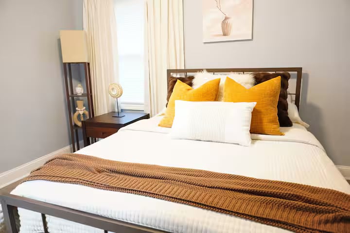 חדר שינה שקט עם מצעים לבנים נקיים, כריות חמות ואור טבעי רך. המיטה מרגישה מזמינה ומאוזנת היטב, עם תאורה ומשטחים בקרבת מקום לנוחות לילית.
