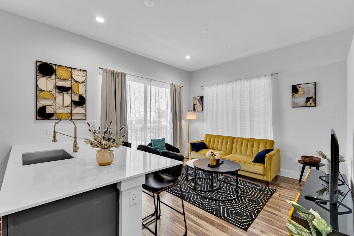 Svetlý otvorený obytný priestor s moderným kuchynským ostrovom, žltou pohovkou, abstraktným umeleckým dielom na stene a prirodzeným svetlom z veľkých okien.