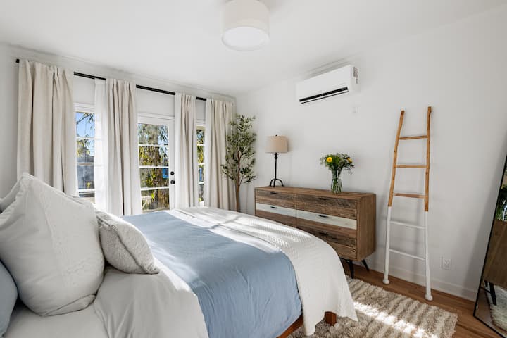 Dhomë gjumi komode me shtroja të bardha dhe blu, jastëkë që përputhen dhe gjelbërim të freskët.