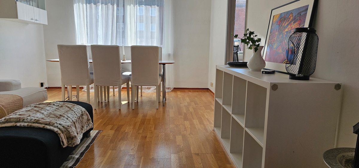 A bright living space features a dining table surrounded by four light-colored chairs. Large windows allow natural light to fill the room, illuminating the hardwood floor. A comfortable sofa with a throw lies in the foreground, while a shelving unit displays decorative items.