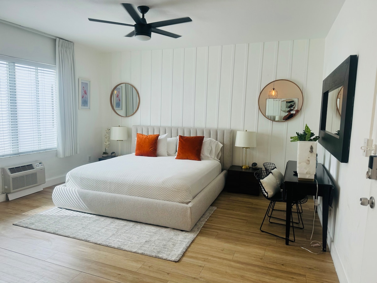 A spacious sleeping area is highlighted by a king size bed with a light-colored duvet and orange accent pillows. Two round mirrors reflect natural light from the window. A small desk and chair provide a workspace beside a low cabinet, enhancing the room's elegant decor.
